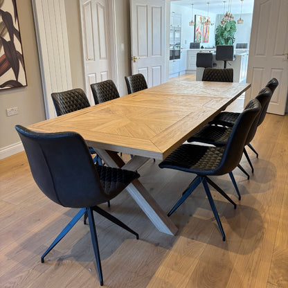 A modern solid oak dining table with a black powder-coated steel frame, surrounded by black chairs with upholstered seats. The table is extendable and set in a room with wooden flooring and a light-colored wall.